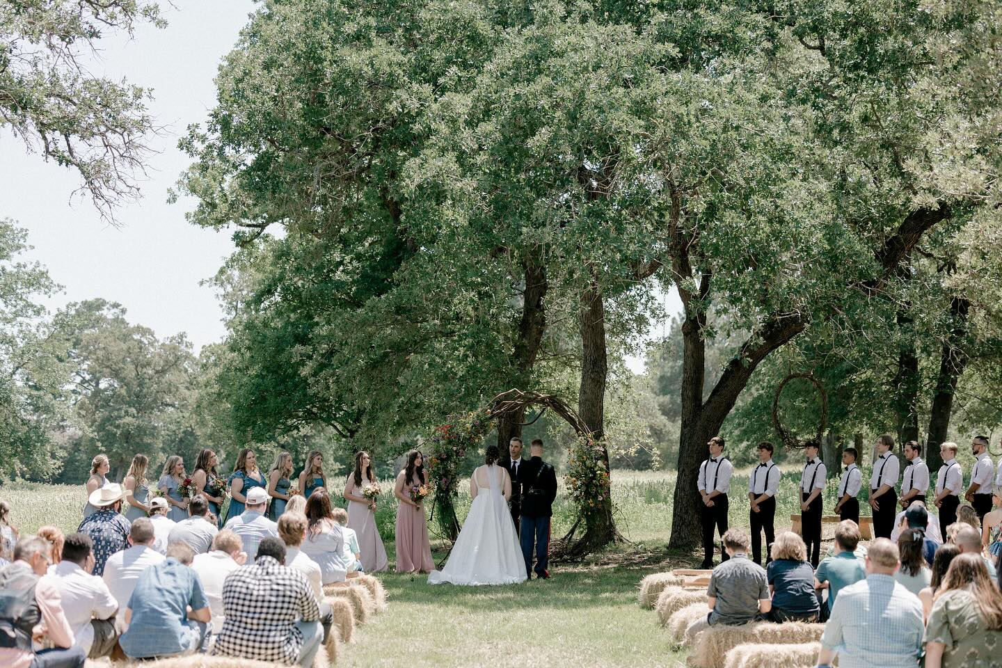 Outdoor wedding ceremony under heritage oaks with floral arbor and hay bale seating