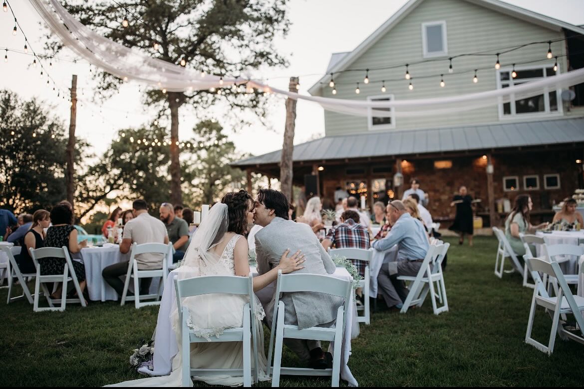 Newlyweds sharing a kiss at outdoor reception with string lights and farmhouse venue